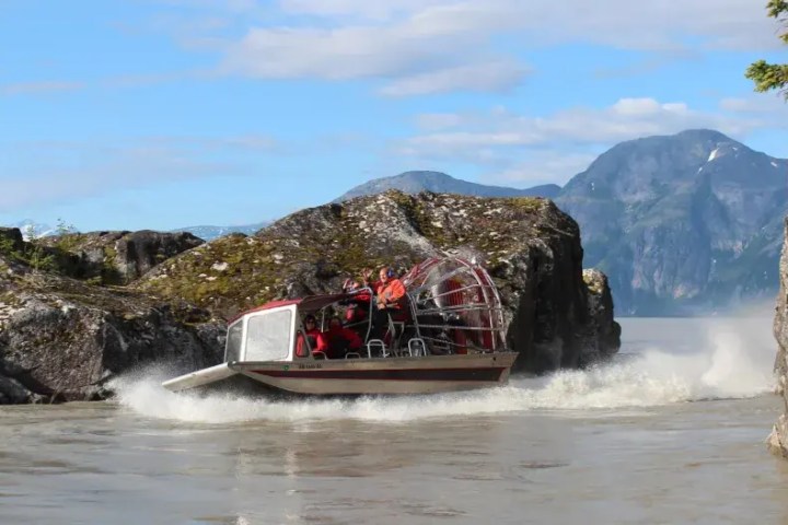 Airboat navigating between rocky cliffs on a river with mountains in the background.
