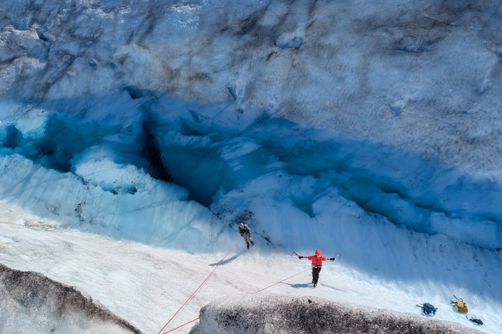 Person in red jacket ice climbing on a glacier with deep blue crevasses and ropes.