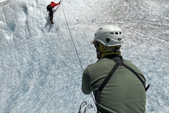 Two climbers ice climbing, one ascending, one belaying, on a steep icy slope under a clear blue sky.