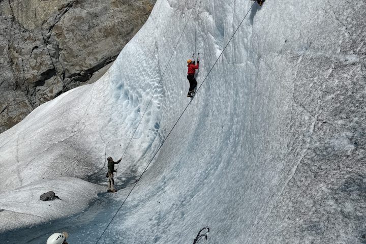 Two climbers scaling an ice wall with ropes on a glacier, wearing helmets and using ice axes.