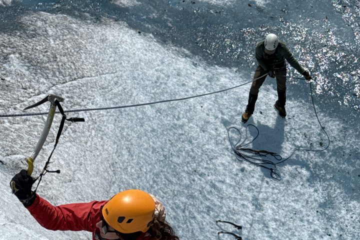 Climber in red jacket and orange helmet scaling icy slope with another holding rope.