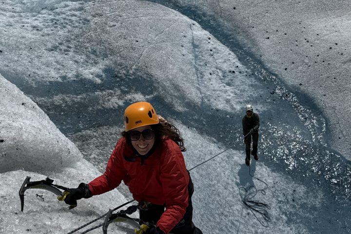Person climbs icy slope with ice axes, another person belaying below on glacier terrain.